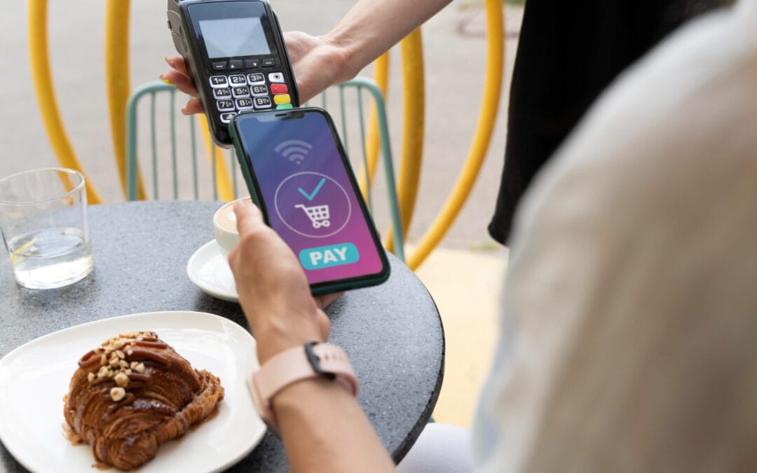 Person using a smartphone to make a contactless payment at an outdoor table with a croissant and a glass of water.