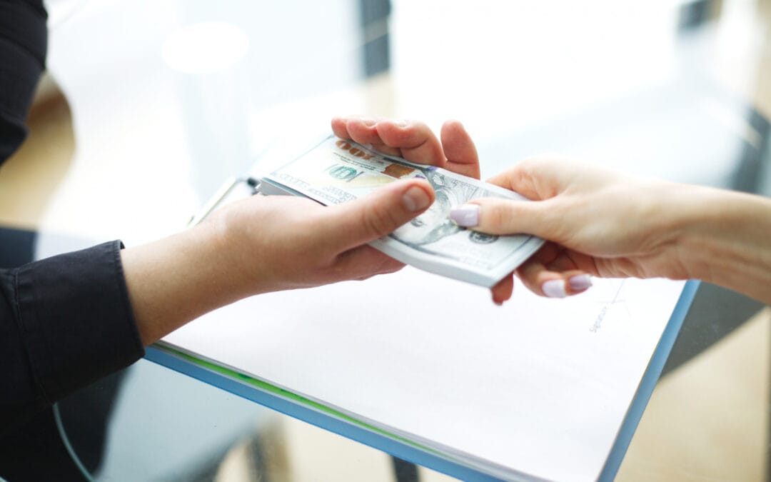 One person hands a stack of U.S. $100 bills to another over a desk with documents.